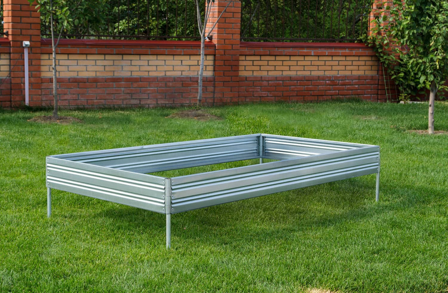 Metal raised Planta garden bed - 1 tier on grass with a brick wall and trees in the background